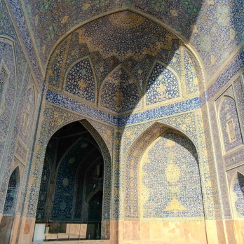 The domed ceiling of a prayer hall at Imam Khomeini Mosque, half bathed in sunset light and half in shadow. Detailed turquoise and golden tiles highlight the mastery of Persian mosaic art