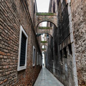 A narrow alley beside the Palazzo delle Prigioni in Venice, lined with tall brick walls and overhead arches that connect the buildings on either side. The historic architecture and the intimate passageway evoke the timeless charm of the city’s hidden corners