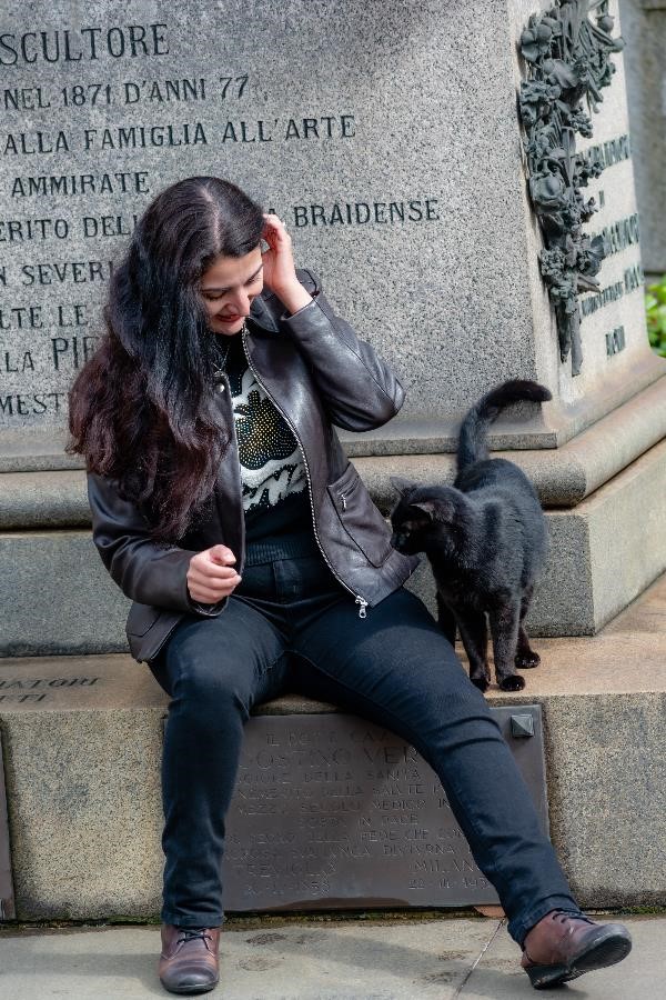 A playful black cat pacing around a woman sitting on a stone platform