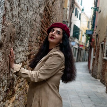 A beautiful woman wearing a small red hat, with striking dark eyes and long flowing hair, stands by the brick wall of Palazzo delle Prigioni in Venice. Her hands rest gently on the wall as she gazes up at the sky, lost in thought. The scene captures a quiet, emotional moment against the backdrop of historic architecture