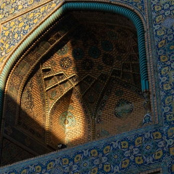 Golden sunset light illuminates the walls of Shah Mosque, enhancing the intricate floral and arabesque tilework in turquoise, blue, and gold. The interplay of light and shadow highlights the beauty of Persian design