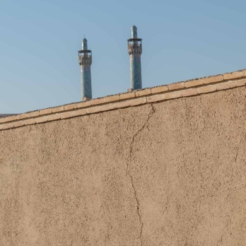 A tall mudbrick wall in a narrow Isfahan alley, behind which two towering minarets of the Imam Khomeini Mosque (Shah Mosque) rise gracefully, blending traditional architecture with the city’s historic charm