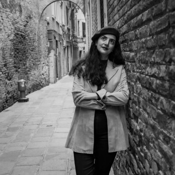 A black-and-white photograph of a Persian woman with long hair, wearing a light velvet coat, leaning against the tall brick wall of Palazzo delle Prigioni in Venice. She gazes upward toward the sky, evoking a sense of quiet reflection and timeless elegance
