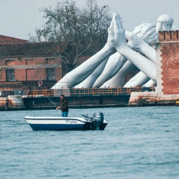 An image of the artwork by Lorenzo Quinn along the waterfront in Venice, with a fishing boat visible on the water in front of it
